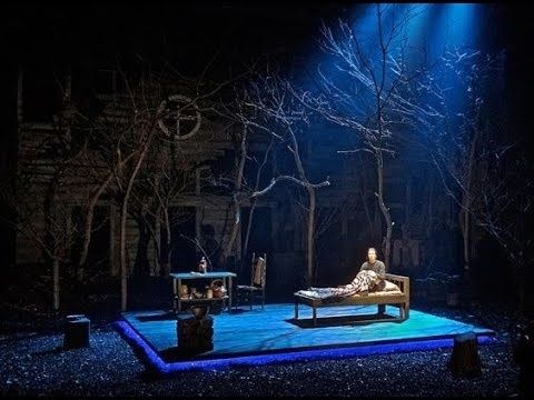 Teaterscene med blått scenelys, nakne trær og en person på en seng